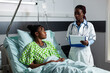 © DC Studio - Doctor of african american ethnicity consulting young patient about healing treatment process in hospital ward. Specialist woman talking to girl sitting in bed with medical equipment