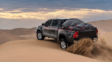 A Black Pickup Truck Is Going Up From A Sand Dune And Splashing Sands On Air And Around In Dasht E Lut Or Sahara Desert With Cloudy Sky