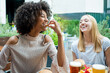 © MandriaPix - two young women having fun while eating snacks at restaurant, group of multiethnic people tasting junk food, diversity and inclusion concept of multicultural lifestyle