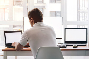 Wall Mural - Man sitting at workplace with two laptops and monitor near the window. Remote work from home