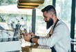 Handsome Barista Using Digital Tablet in a Cafe.
 
Serious bartender with a beard standing on the bar counter and reading online order on a digital tablet while working in the restaurant.
