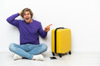 © luismolinero - Young blonde man with suitcase sitting on the floor surprised and pointing finger to the side