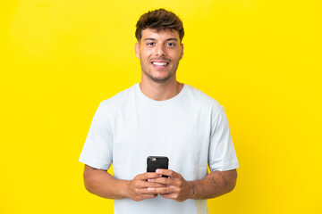 Wall Mural - Young caucasian handsome man isolated on yellow background looking at the camera and smiling while using the mobile