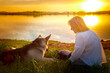Portrait of middle-aged woman near lake or river and her dog german shepherd during beautiful yellow sunset with sun above the horizon and reflection in the water