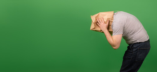An unrecognizable man with a paper bag on his head grabbed his head against a green background. Place for your text. mental health, greenwashing, disinformation concept