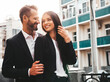 © halayalex - Beautiful fashion woman and her handsome boyfriend in suit. Sexy smiling brunette model in black evening dress. Fashionable couple posing in the street at sunset. Brutal man and his female outdoors