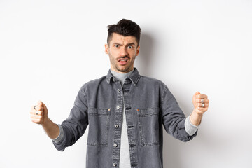 Skeptical young man showing big size object and frowning doubtful, standing reluctant on white background