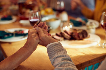 Wall Mural - Close-up of senior couple holds hands while blessing food during Thanksgiving meal with family at dining table.