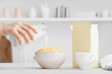 Woman pouring milk from bottle into bowl with corn flakes on table in kitchen