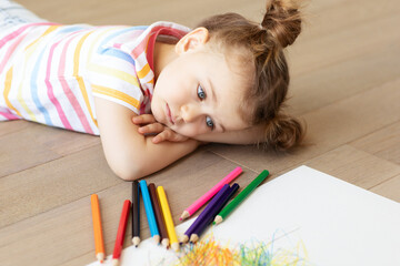 Wall Mural - Sad tired frustrated bored stressful child lies on wooden floor with white sheet of paper and colored pencils. Learning difficulties, education concept. Family relationship. Misses home