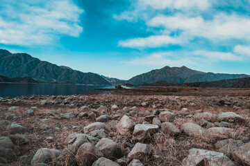 Canvas Print - Mesmerizing view of a rocky field, calm lake with hills on the background under a cloudy sky