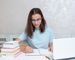 © Alena - A smiling girl student sitting at a table with a laptop writes a task in a notebook. Girl student studies at home.