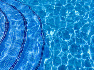 Curved steps into swimming pool, view from above through water on blue mosaic tiles. Waves on water surface. Distortion. Refraction