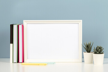 White blank wooden mock up frame on white desk with paper notebook, books, flowers at home