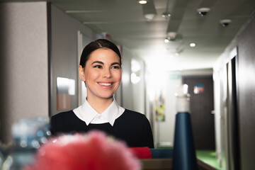 cheerful young maid near blurred housekeeping cart