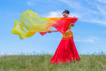 Wall Mural - Young woman in colorful dress is dancing fascinating oriental dance with bright yellow-red fans on green meadow against blue summer sky. Dancing in open air.