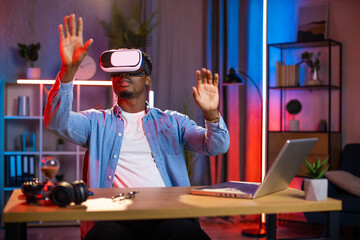 African american man sitting at home office and using VR glasses and laptop for remote work. Black male freelancer touching virtual screen with fingers.