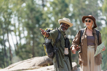 Smiling african american hiker pointing with finger near wife with map in forest.