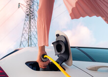 Female Hands Plugging In The Charger Into A Socket Of White Electric Car At A Charging Station Near An Electric Power Plant