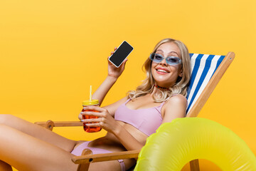 happy woman in sunglasses and swimsuit sitting in deck chair with cocktail and talking on smartphone isolated on yellow.