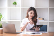 © Daenin - Portrait of a smiling woman working at home in casual clothes. She sat at a large brown wooden table holding a tablet and looked at the laptop and documents in front of her.