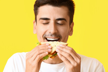 Wall Mural - Young man eating tasty sandwich on color background