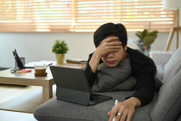 Overworked Asian man working with computer tablet on sofa.