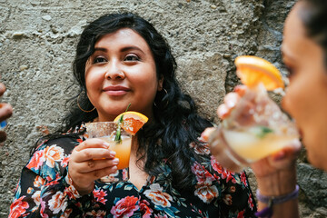 Smiling young woman enjoying cocktail with female friend against wall during weekend garden party in backyard