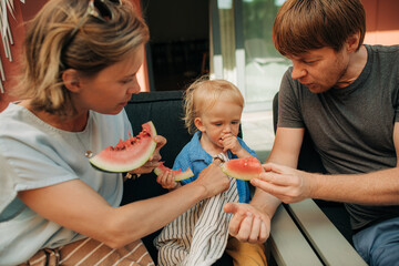 Wall Mural - Family with little girl eating watermelon together. Mid adult parents sitting in chairs and feeding their daughter with fresh fruit. Childcare, family meal concept