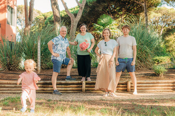 Wall Mural - Portrait of happy family and little child outdoors. Mid adult and mature couples posing outdoors, toddler girl walking nearby. Multi generation family concept