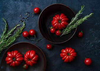 Ripe red tomatoes lie on a black plate on a black background with rosemary dishes, top view.