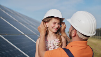 Wall Mural - Father with little girl at solar power plant. The father talks about solar energy. The concept of green energy will save the planet for children. The father puts a protective helmet on the girl's head