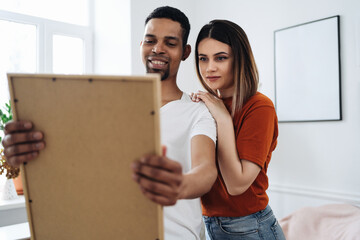 Excited african young husband and caucasian wife hang frame picture on wall happy settling together moving to new house, overjoyed biracial couple decorating own home relocating, ownership concept