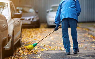 Street cleaner sweeping yellow fallen leaves outdoors, autumn seasonal cleaning work. Cleaner removes leafs, janitor with broomstick remove yellow leaves from road. Autumn street sweeper work.