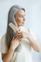 Wall Mural - Pretty middle aged Asian person brushes straight grey hair posing on light background in studio. Mature beauty lifestyle