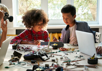 Asian school kid boy helping African child girl student using tablet computer studying programming robots learning making robotic cars sitting at table at STEM education coding engineering class.