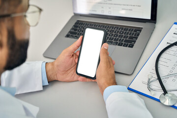 Male doctor gp physician holding mobile cell phone in hands looking at white blank empty mockup smartphone screen working in hospital. Ehealth apps technology mock up concept. Over shoulder view