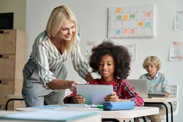 Happy female teacher tutor helping African American junior school kid girl student using digital tablet computer education program learning app technology during elementary class lesson in classroom.