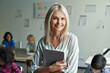 © insta_photos - Happy smiling middle aged woman elementary or junior school female teacher standing in classroom looking at camera holding digital tablet teaching children students during class. Portrait
