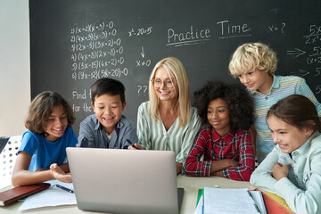 Happy diverse junior school children students gathered at teacher table looking at laptop computer using online software learning web education technology studying together at math class in classroom.