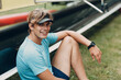 Sportsman single scull man rower portrait sitting relaxing after training competition against background with boats.