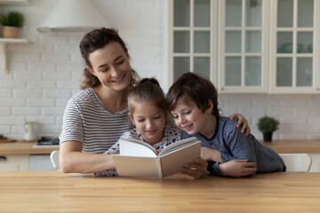 Happy mom and two children enjoying book story in kitchen. Mother entertaining kids at home, teaching boy and girl to read, improving imagination, interest to literature. Family smart activity