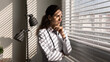 © fizkes - Smiling thoughtful female doctor standing by window in office, looking away, thinking of future vision, successful medical career growth, promotion, success. Portrait with shutter shadows