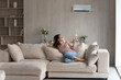© fizkes - Relaxed young hispanic female homeowner sitting on huge comfortable couch, turning on air conditioner with remote controller, switching on cooler system, setting comfortable temperature in living room