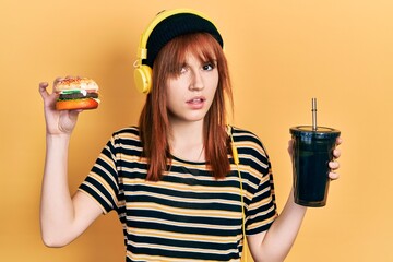 Canvas Print - Redhead young woman eating a burger and drinking soda wearing headphones clueless and confused expression. doubt concept.