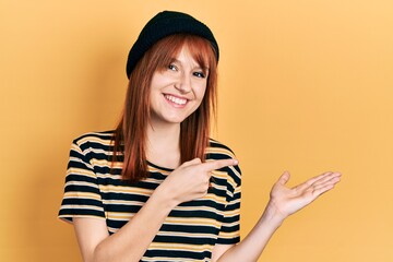 Sticker - Redhead young woman wearing wool cap amazed and smiling to the camera while presenting with hand and pointing with finger.