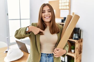 Wall Mural - Young brunette woman holding paper blueprints at the office pointing finger to one self smiling happy and proud