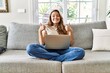 Beautiful young brunette woman sitting on the sofa using computer laptop at home success sign doing positive gesture with hand, thumbs up smiling and happy. cheerful expression and winner gesture.