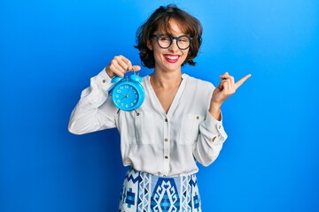 Sticker - Young brunette woman holding alarm clock smiling happy pointing with hand and finger to the side