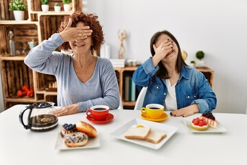 Poster - Family of mother and down syndrome daughter sitting at home eating breakfast smiling and laughing with hand on face covering eyes for surprise. blind concept.